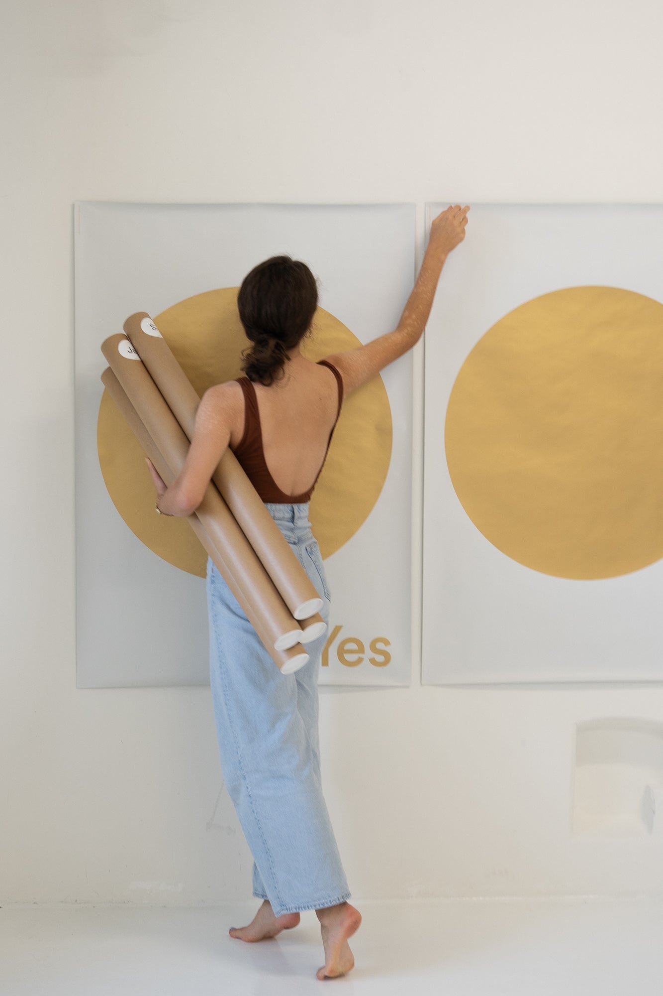 A woman hanging original plakat ja oui si posters on a wall, holding rolled prints with gold circles.