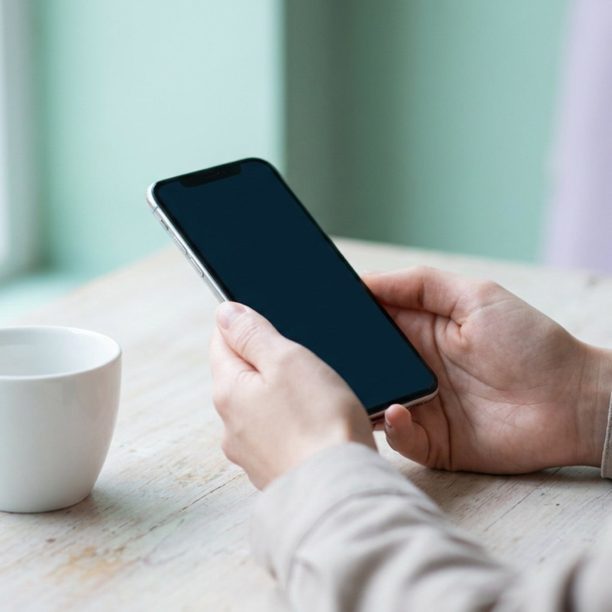 Zwei Hände halten ein Telefon über einem hellen Tisch, daneben eine weiße Tasse