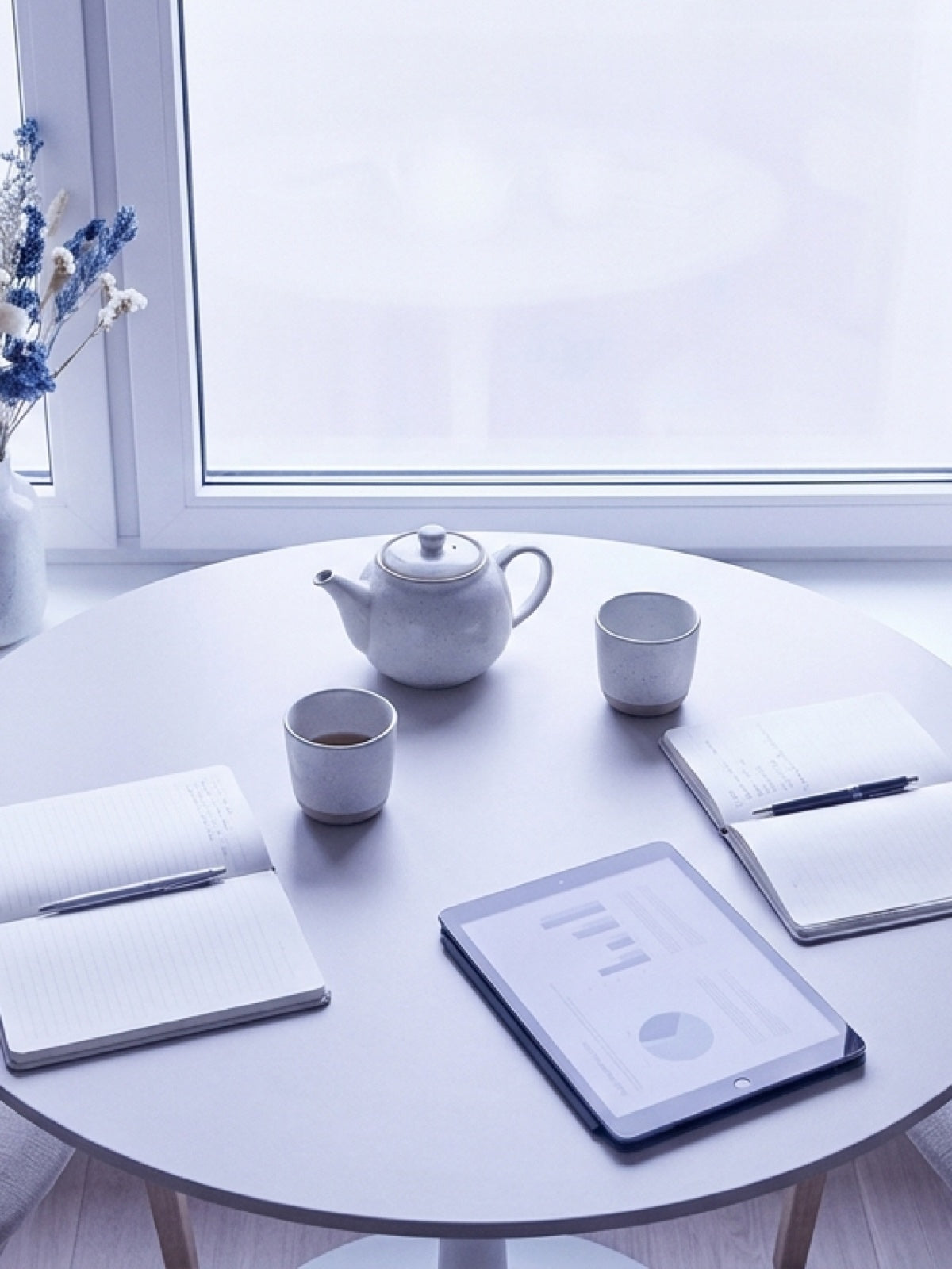 Runder Tisch am Fenster mit Teekanne, zwei Tassen, zwei Notizbüchern und einem Tablet